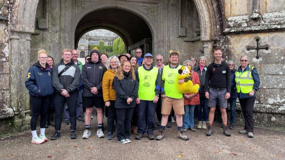 A group of about 20 people is gathered in front of a stone archway that forms part of an old, weathered building. They stand on a gravel path, dressed in casual outdoor clothing, with some wearing bright fluorescent vests. Among them, James Dundon is holding a plush toy bear about 2ft high.