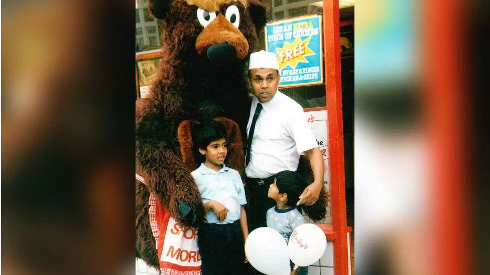 A man, two children, and a person in a bear costume stand outside a Morley’s Fried Chicken shop.
