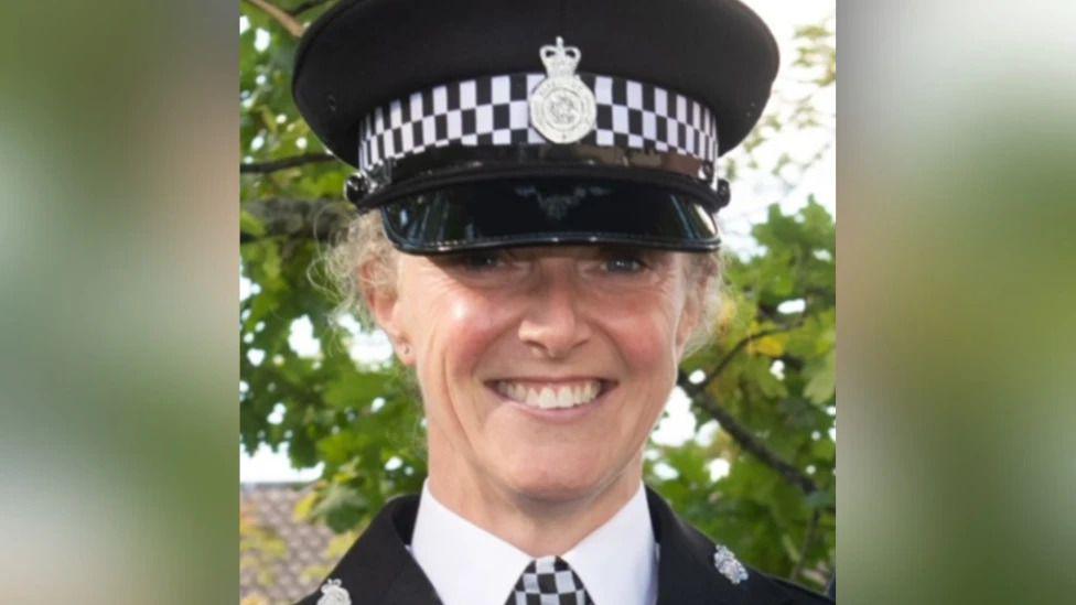 A female police officer is smiling at the camera, in full uniform. Her hair is blonde, and she's wearing a police hat. There are tree branches visible in the background, surrounded by green leaves.