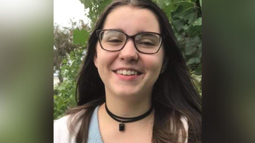 Bernadette Walker is smiling at the camera. She has long, dark brown hair and is wearing dark-rimmed spectacles. She has a leather necklace on and a blue top and white jacket