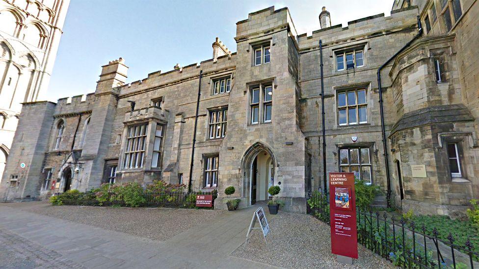 The outside of the Visitor and Learning Centre, close to the main entrance of Peterborough Cathedral. It is an old, grand stone building like a castle, with green foliage outside and a red plinth advertising the centre.