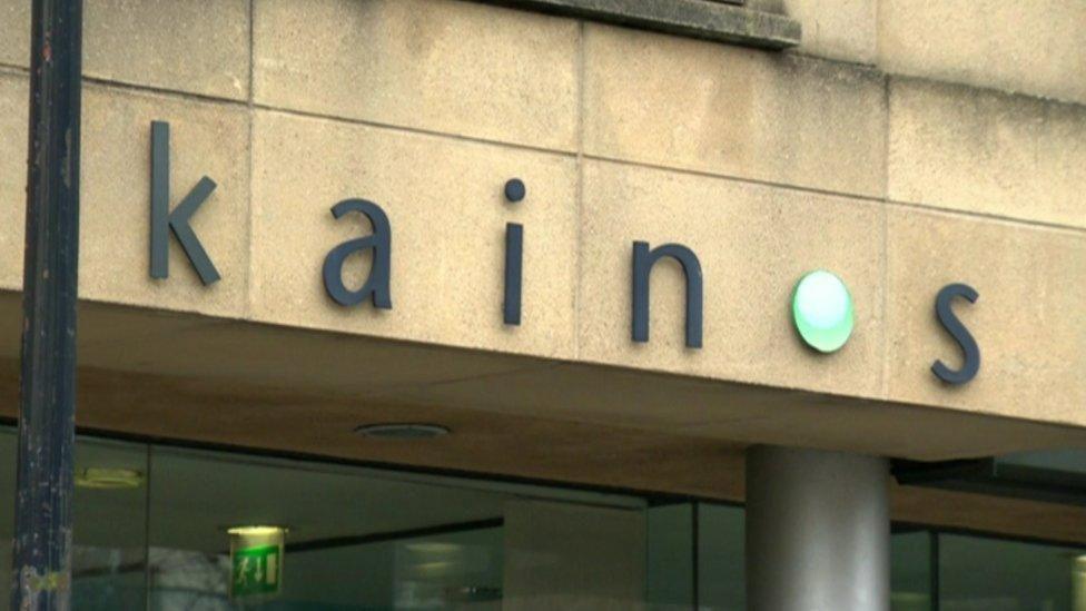 A shot of the exterior of Kainos's building in Belfast. The logo Kainos written in lower case is in black with a green circle dot replacing the O.