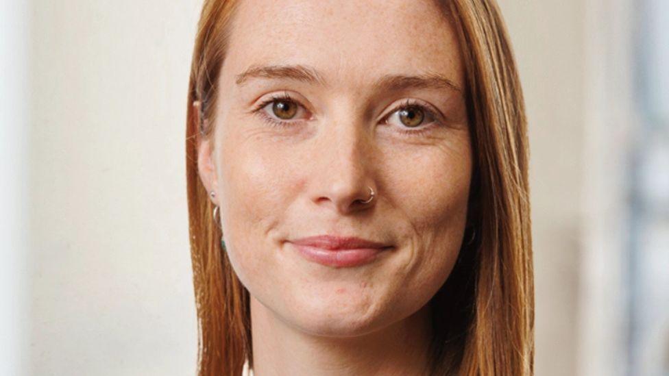 A woman with brown hair and brown eyes looks into the camera. She is wearing a nose ring and earrings.