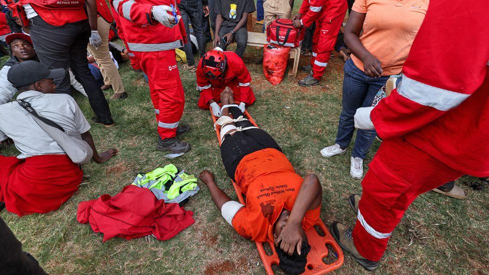 An injured woman in an orange T-shirt lies on a stretcher on the grass surrounded by Red Cross workers at a stadium in Nairobi.
