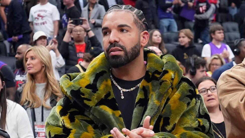 Drake pictured in a furry camouflage jacket and black t-shirt and silver chain.