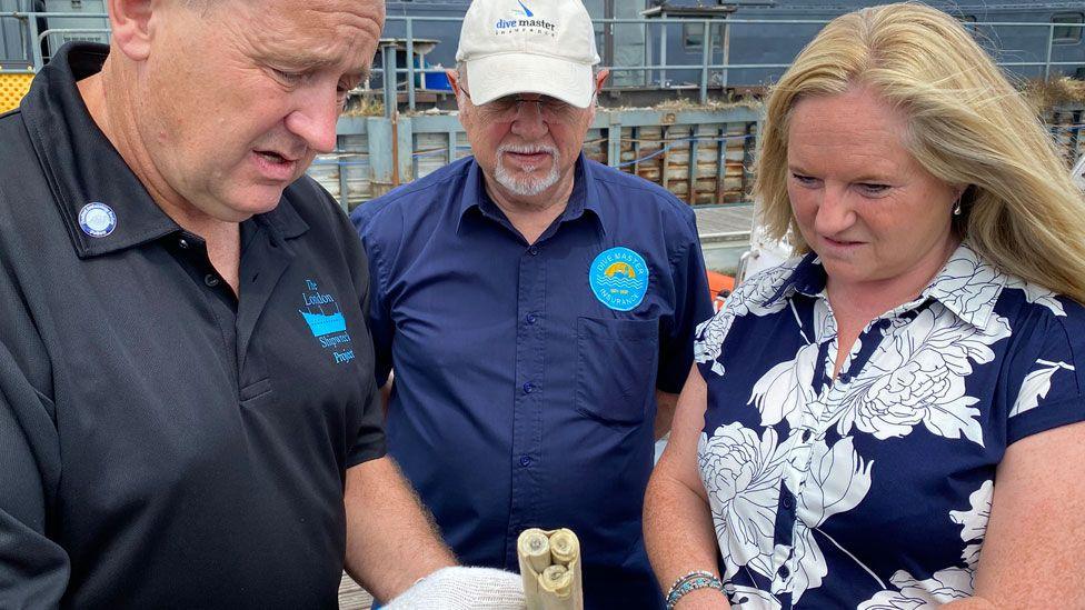 Steve Ellis showing Bob Archell and Helen Gray a 17th Century candle. He is on the left, dressed in a black polo-shirt with a turquoise logo on his left chest saying the London Shipwreck Project. Bob Archell is in the middle, wearing a navy blue short-sleeved shirt and white cap and has a white goatee beard, while Helen Gray is on the right, wearing a navy dress with large white flowers and has long blonde hair. They are all looking down at the candle and are standing on a boat's deck.
