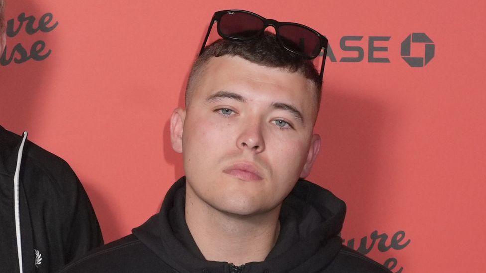 Close shot of a dark haired young man with short brown hair, a black hooded jacket and dark sunglasses on top his head