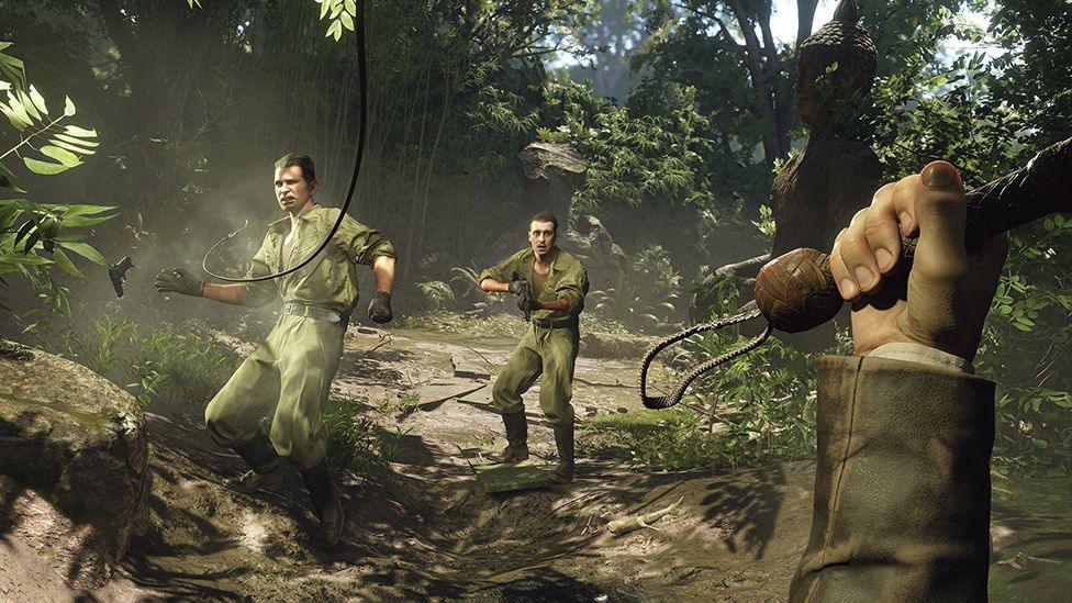 Screenshot of a jungle scene taken from a first-person perspective shows the player's hand holding the handle of a bullwhip. The end of it can be seen striking one of two enemy characters dressed in combat fatigues about 10 feet away from the player. The one who's just been hit is recoiling and a pistol can be seen falling out of his hand. The other looks on with a panicked expression as he readies his own weapon.