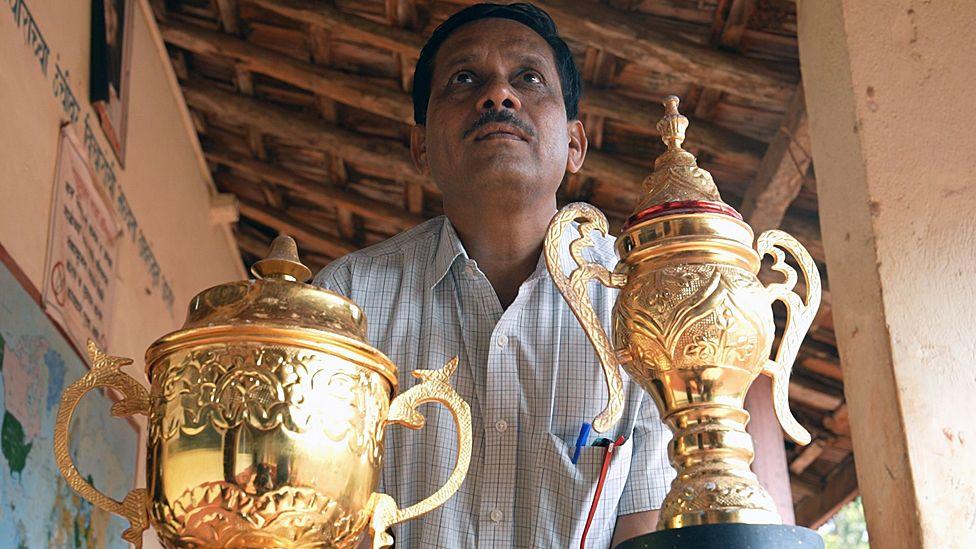 Daji Rajguru in a checked shirt with two large golden trophies in front of him. The trophies are embossed with patterns and are shaped like vases with lids and handles. 