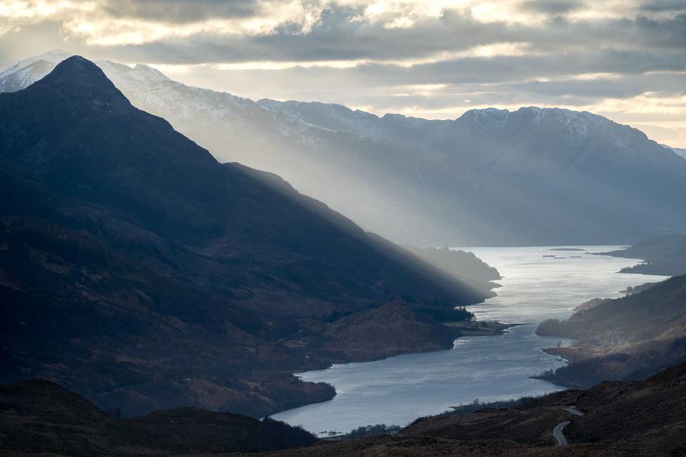 Hillsides, some snow-capped, next to a loch, with evening sunlight, and a small glimpse of a winding road.