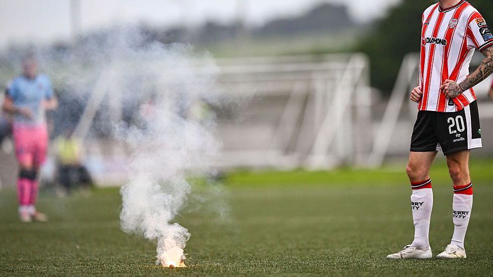 A flare is lying on the pitch and a Derry City player is standing to the side.