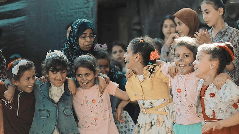 A group of children, dressed in an array of pastel shades, smile and laugh, with their arms around each other's shoulders. 