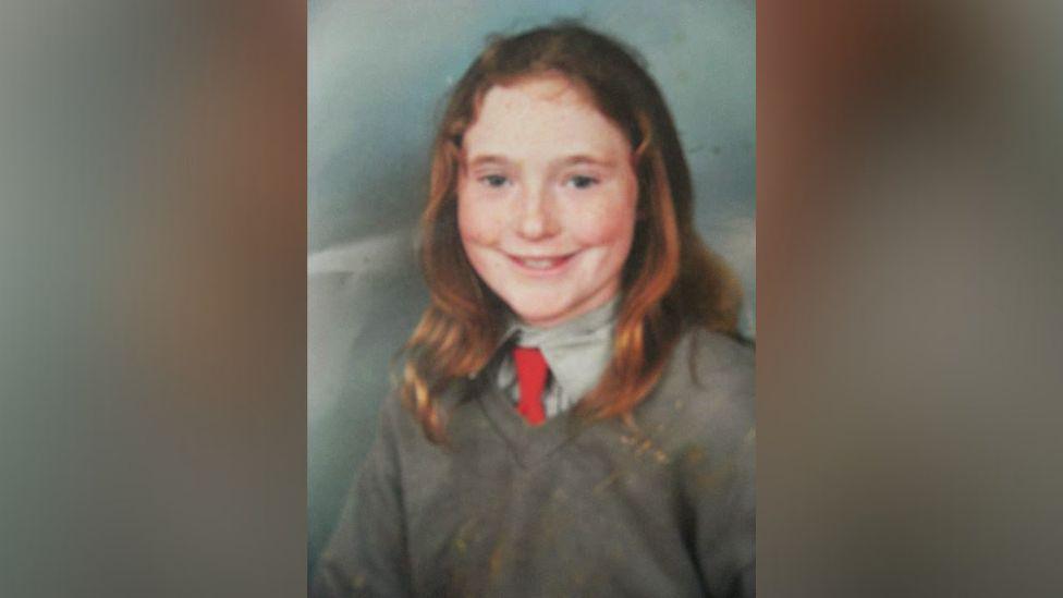 Danielle McLaughlin as a young child in a school photograph. She has long ginger hair and is wearing a grey v neck jumper with a grey shirt and a red tie. 