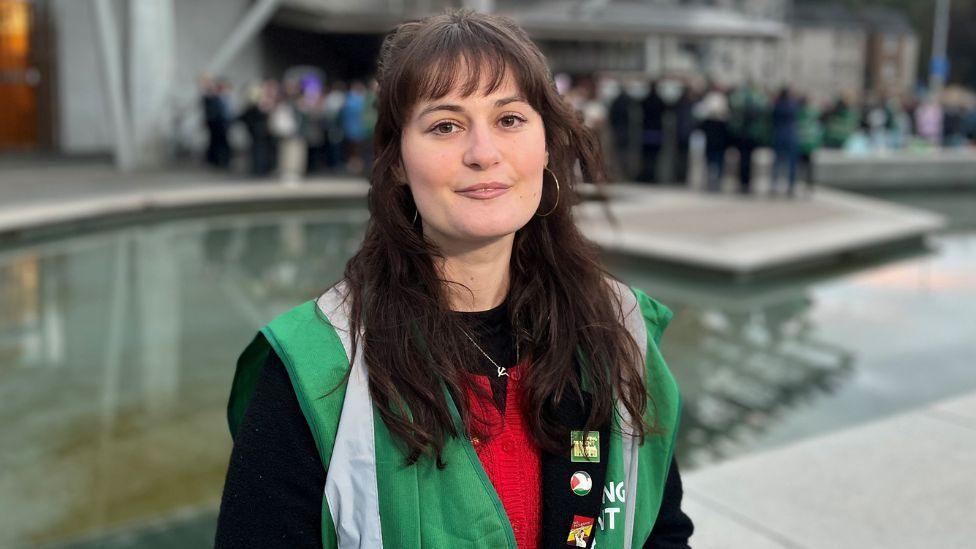 Bianca Lopez, who has long dark hair, looks at the camera while standing outside the Scottish Parliament. She is wearing a green vest over a red and black top. There is a crowd of people behind her, standing at the far side of a water feature. 