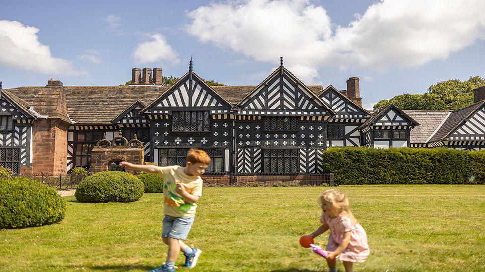 A young boy with red hair, yellow t-shirt and blue denim shorts plays on the lawn in front of Speke Hall alongside a young girl in a pink dress with blond hair holding a ball on a clear day.