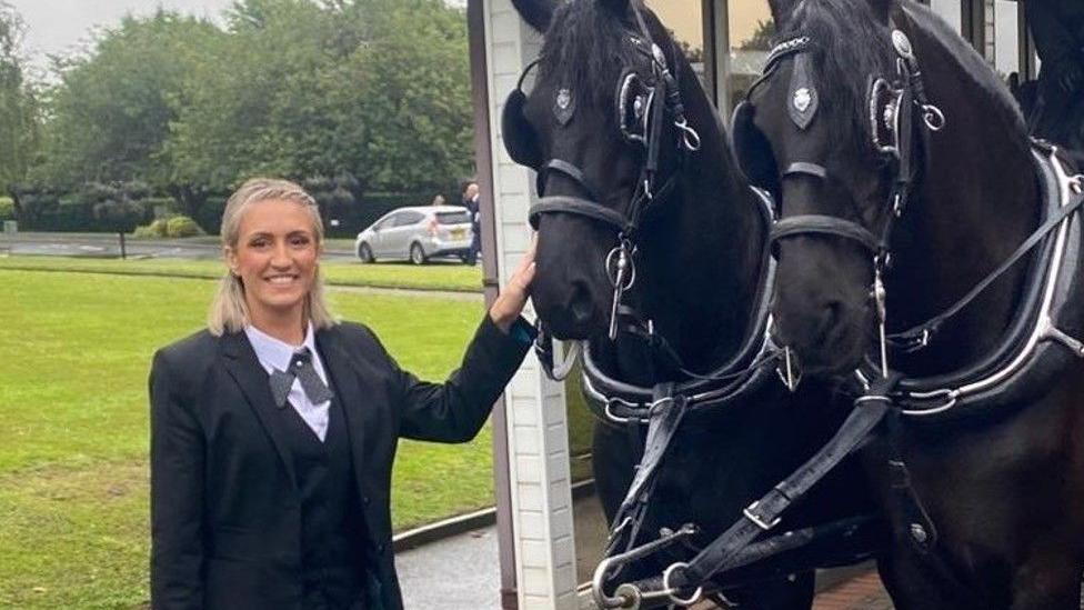 A woman largely wearing black is standing near two black horses.