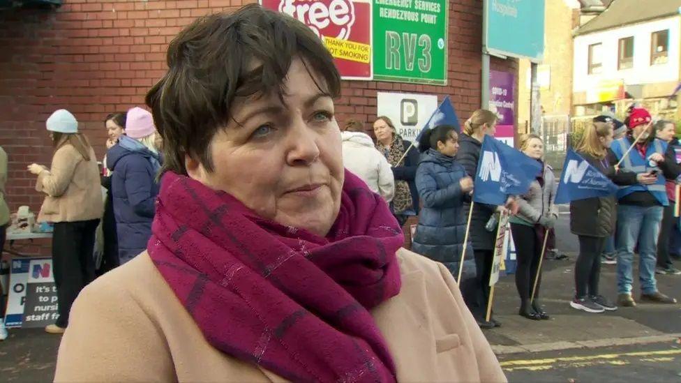 Rita Devlin with short dark hair, wearing a red scarf and beige coat while standing in front of a picket line