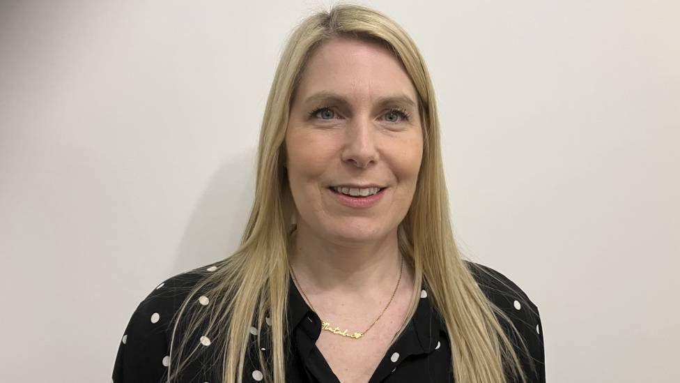 A white smiling woman with long blonde straight hair wearing a black polka dot blouse 