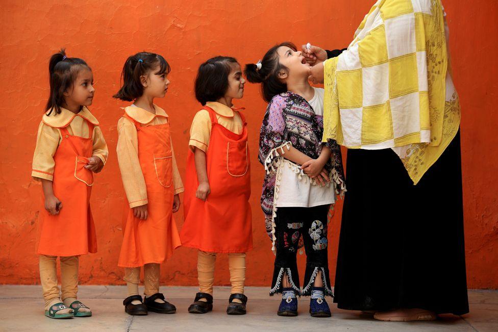 Three children in red pinafore dress uniforms queue behind a child in a black and purple shawl as she receives an oral vaccination in Peshawar, Pakistan, 28 October 2024.  An adult wearing a black dress and a yellow and white check shawl is putting the droplets into the child's mouth.