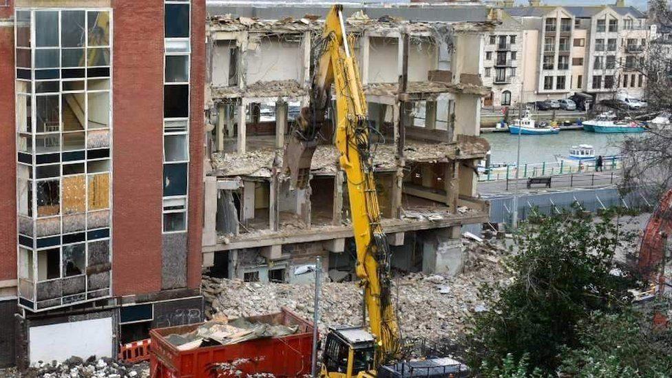 North Quay, a multi-storey office building, being demolished with a yellow digger. In the background, Weymouth Harbour can be seen