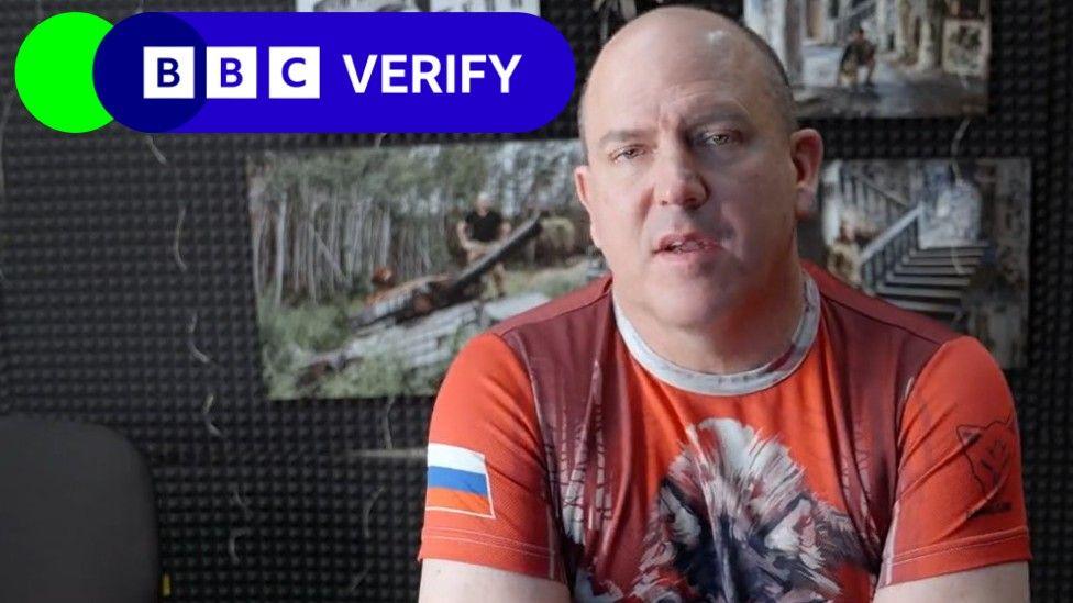 A bald man looking at the camera wearing a t-shirt with a wolf on the front and a Russian flag on the sleeve