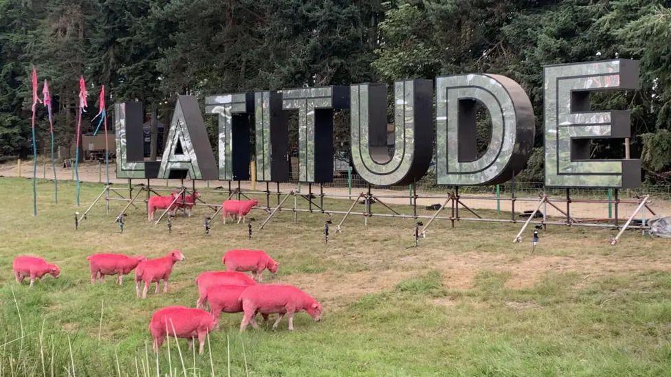 Very tall metal letters spelling Latitude with dark green trees behind them and a flock of pink-dyed sheep grazing the grass in front of them