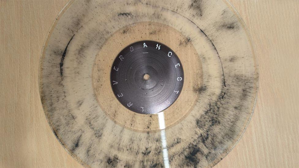 The transparent vinyl record with black coal dust particles embedded in it. It's sitting on a pine coloured table. The centre of the disc is black, with letters in silver that are tricky to read clearly.