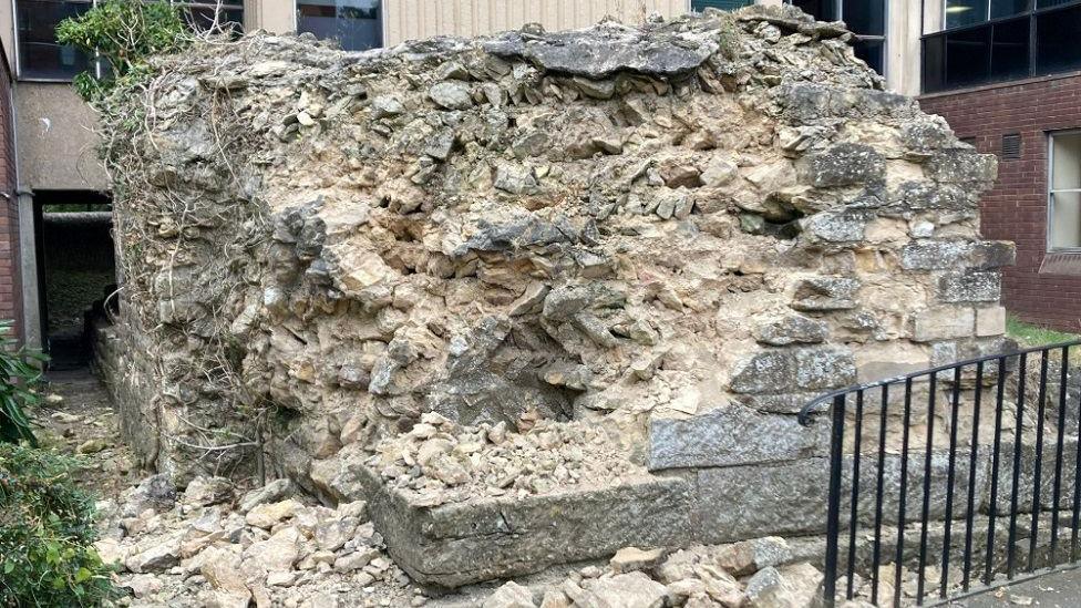 Light-coloured bricks are seen damaged and in a pile of rubble with a black railing and green bushes next to the wall.