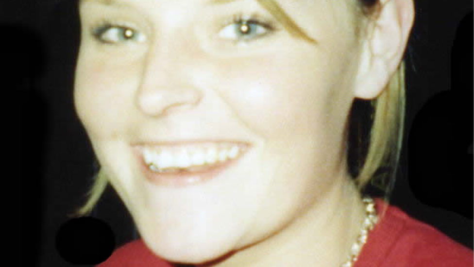 A close up picture of a blonde woman with her hair up. She's smiling. She's wearing a red top, which is just visible, and a necklace.  