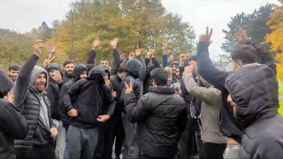 Men mostly wearing black jackets, coats and hoodies appear to stand and jump around, with trees in the background.