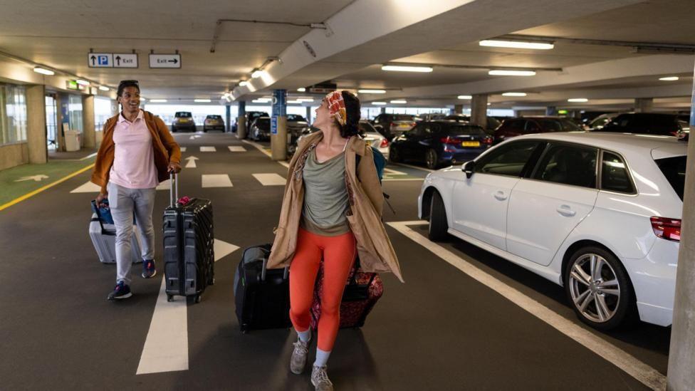 A couple wheeling large suitcases through an indoor car park. They are stood next to a white car.