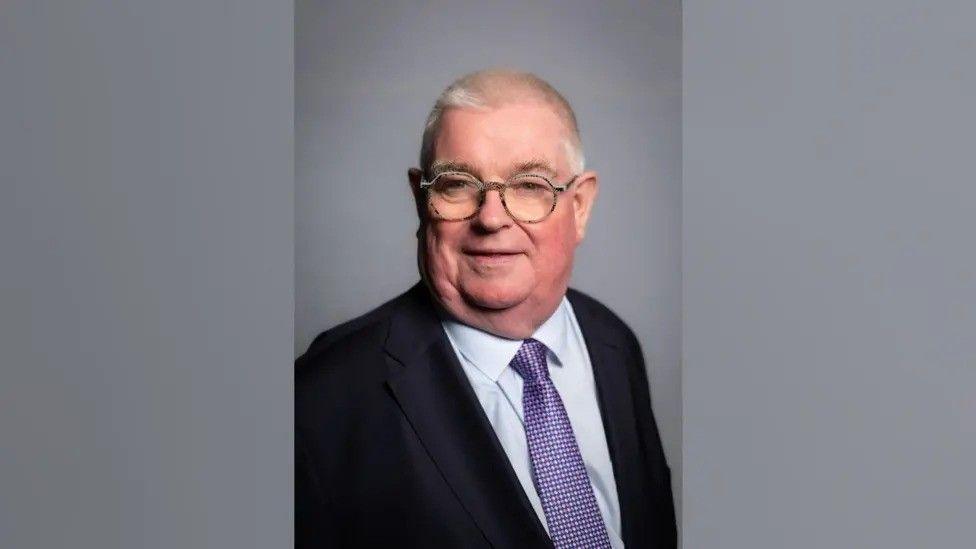 John Tizard with white hair and round glasses. He has a black suit on, with a light blue shirt and a purple spotted tie. It is an official photograph and he is standing in front of a grey background.