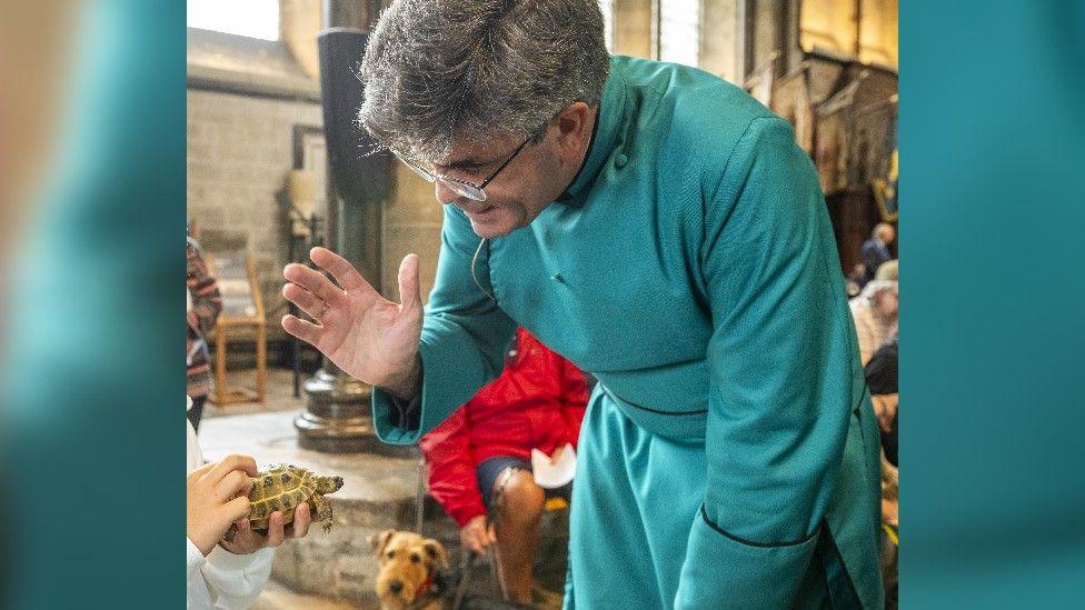 A Dean, dressed in a teal gown and wearing glasses, raises his hand as he blesses a tiny tortoise, being held by a child as a dog watches on.