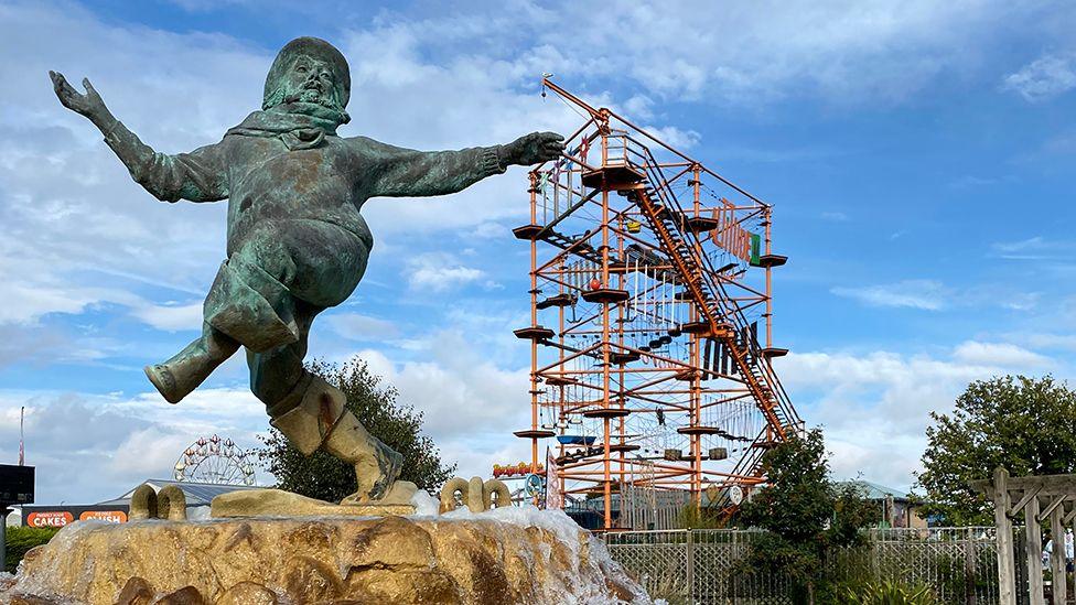 The Jolly Fisherman statue is bronze and covered in green Verdigris. It stands on a stone plinth with water bubbling over. In the background is a large red climbing frame.