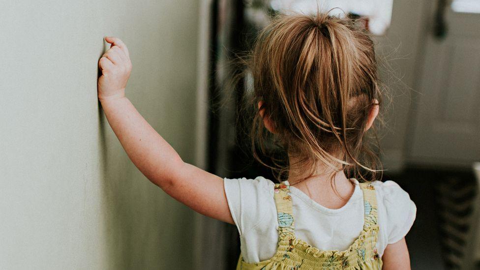 The back of a toddler girl who is walking down some stairs at a house. She has her left arm against the wall to steady herself. She has long blonde hair and is wearing a white T-shirt and dungarees.