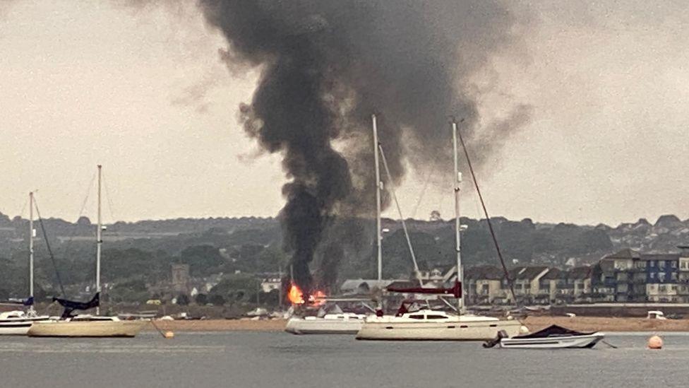 Thick black smoke pours into the sky from a large fire in Exmouth. Boats are on the water in front of the fire. Buildings are in the background.