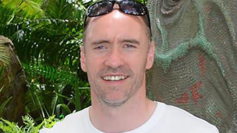 Paul Doyle has short dark greying hair with sunglasses on his head and is wearing a white T-shirt.