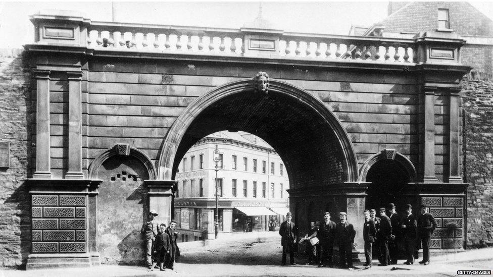 A early 20th century black and white photograph of Ferryquay Gate, one of the gates of Derry's walls. An archway can be seen in the old ramparts, through which shops can be seen., A number of men stand at the foot of the gate, as the walls reach up above them