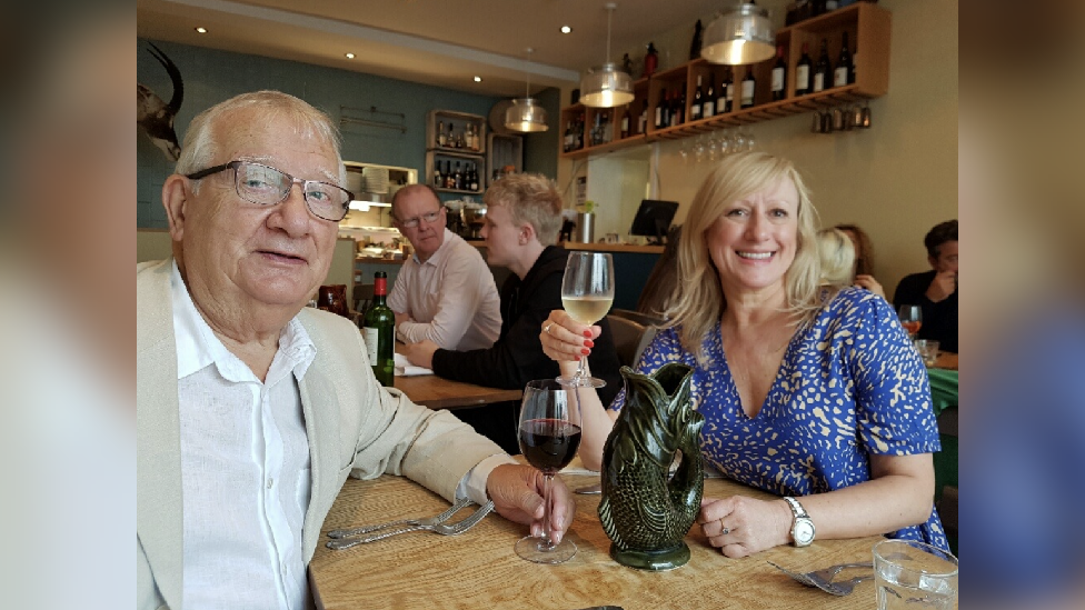 Anna-Louise Marsh-Rees in blue and white printed dress is sat across the table from her dad who is wearing a cream jacket and white shirt. They are both looking at the camera. He has a glass of red wine on the table and is holding it with his left hand. She has a glass of white and it is in her right hand. They are in a restaurant with people around them. 