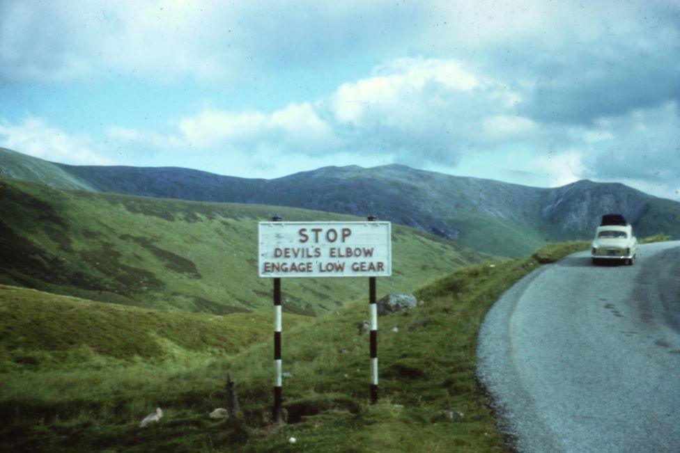 Sign in old photo which says 'Stop: Devil's Elbow engage low gear' on a winding hillside road with an old car in the background.