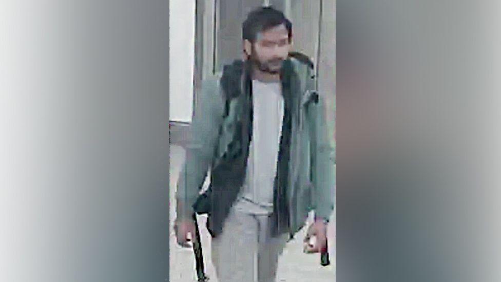 CCTV of Pankaj Lamba wearing a green coat, white top and grey pants. He looks to have a backpack and is walking. 