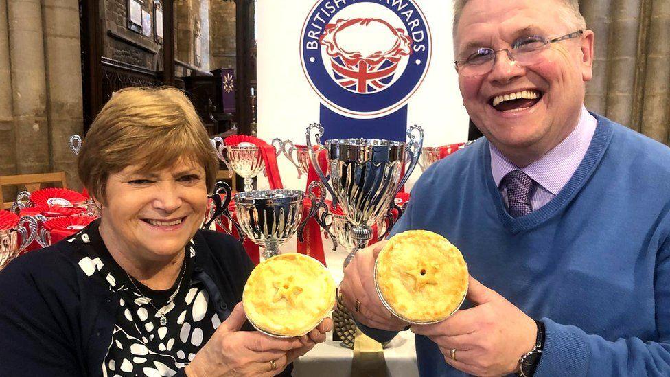 Christine has short brown hair and is smiling. She is wearing a black cardigan and has a black and white spotted blouse on underneath. Paul is laughing at the camera. He is wearing a blue jumper over a pale purple shirt and tie. He is wearing glasses. They are each holding a pie up to the camera.