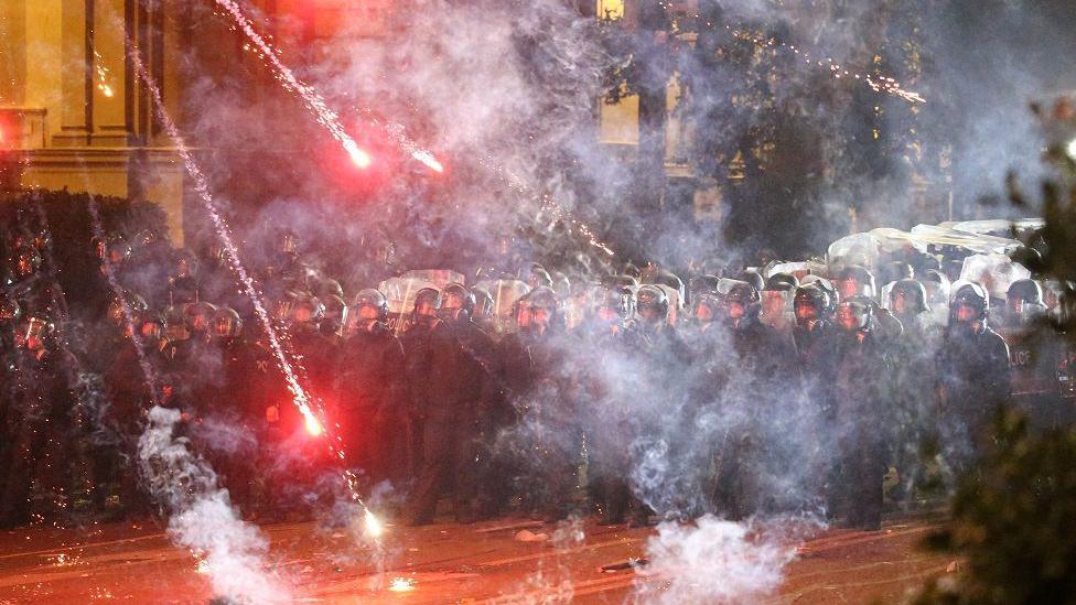 Fireworks land near a barricade of police, who are in riot gear and holding up shields
