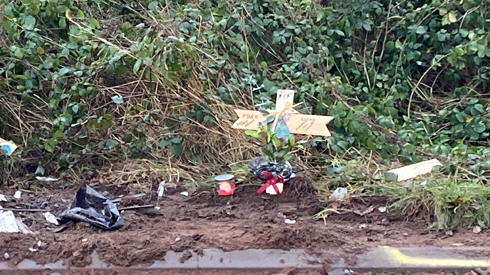 Memorials, including a wooden cross next to a hedge by the side of the road where the person died