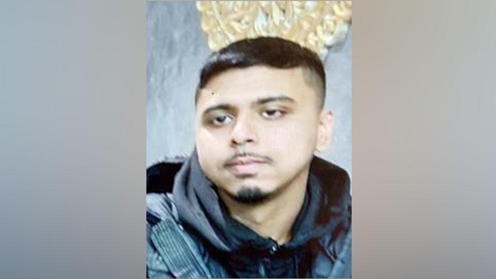 Yusef pictured indoors in front of a grey wall and cream pattered ornament. He is wearing a dark hoodie and coat over the top, and is staring slightly to his right, not at the camera. He has brown eyes and black hair, and a goatee beard and moustache.