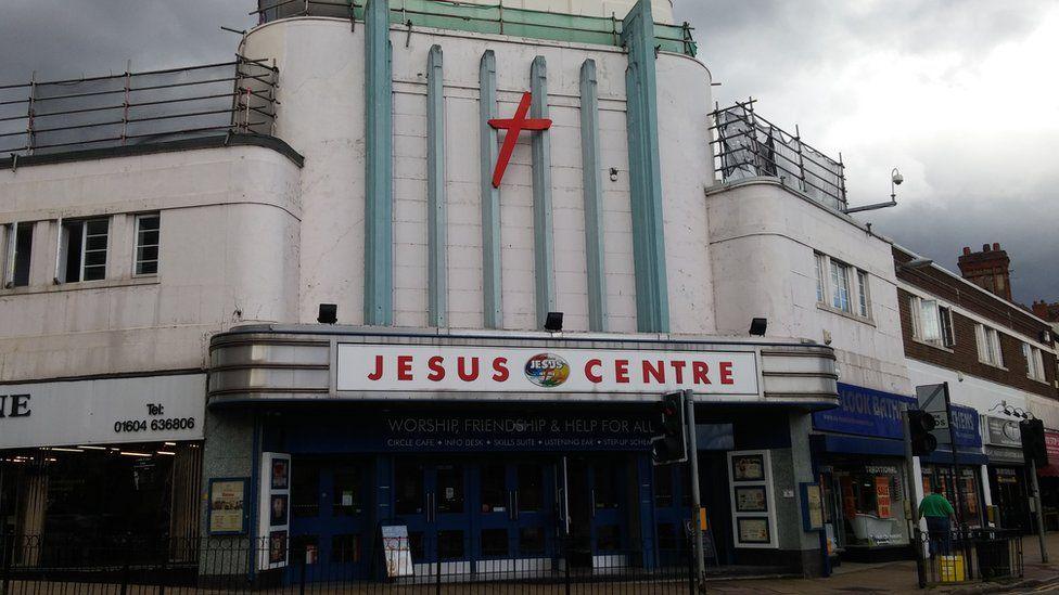 A large white and green building with "Jesus Centre" written across it with a large red crucifix on the outside.