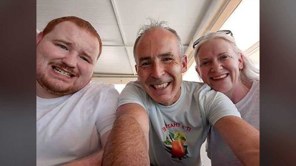 Evan O'Connor and his parents Mark and Louise O'Connor smiling at the camera in a family photo.   Evan has short, ginger hair and a beard.  Mark and Louise both have grey hair.  They are all wearing white t-shirts. 