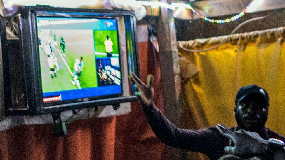 People gesturing at a television screen which is showing a football match - Nigeria 2018