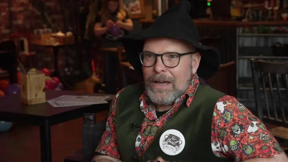 A man is wearing a black wizard's hat, he has glasses and a short beard. He is sitting in a chair wearing a brightly patterned red shirt with a black waistcoat