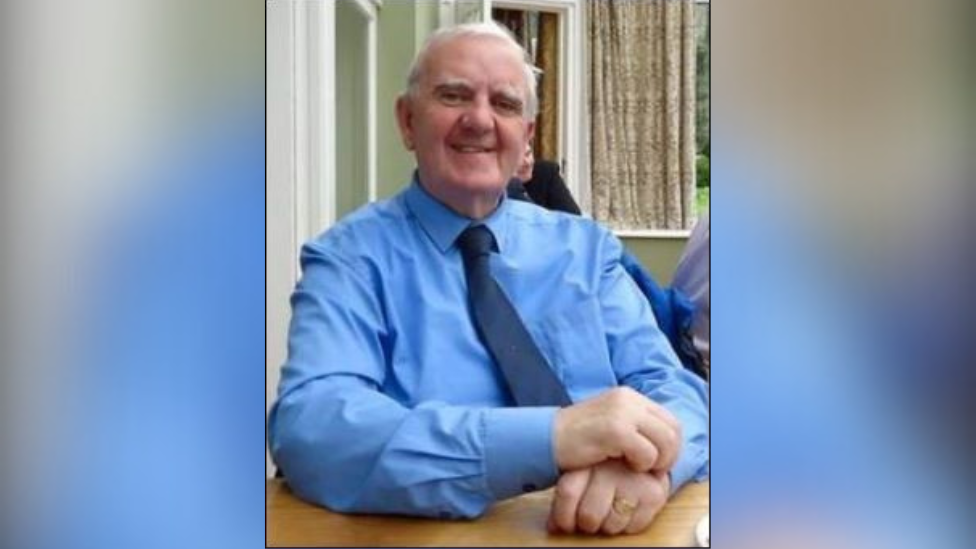 Billy Dickson. He is sitting at a table with his arms placed on the table. He has white hair, wearing a blue shirt and dark blue tie and a gold signet ring. 